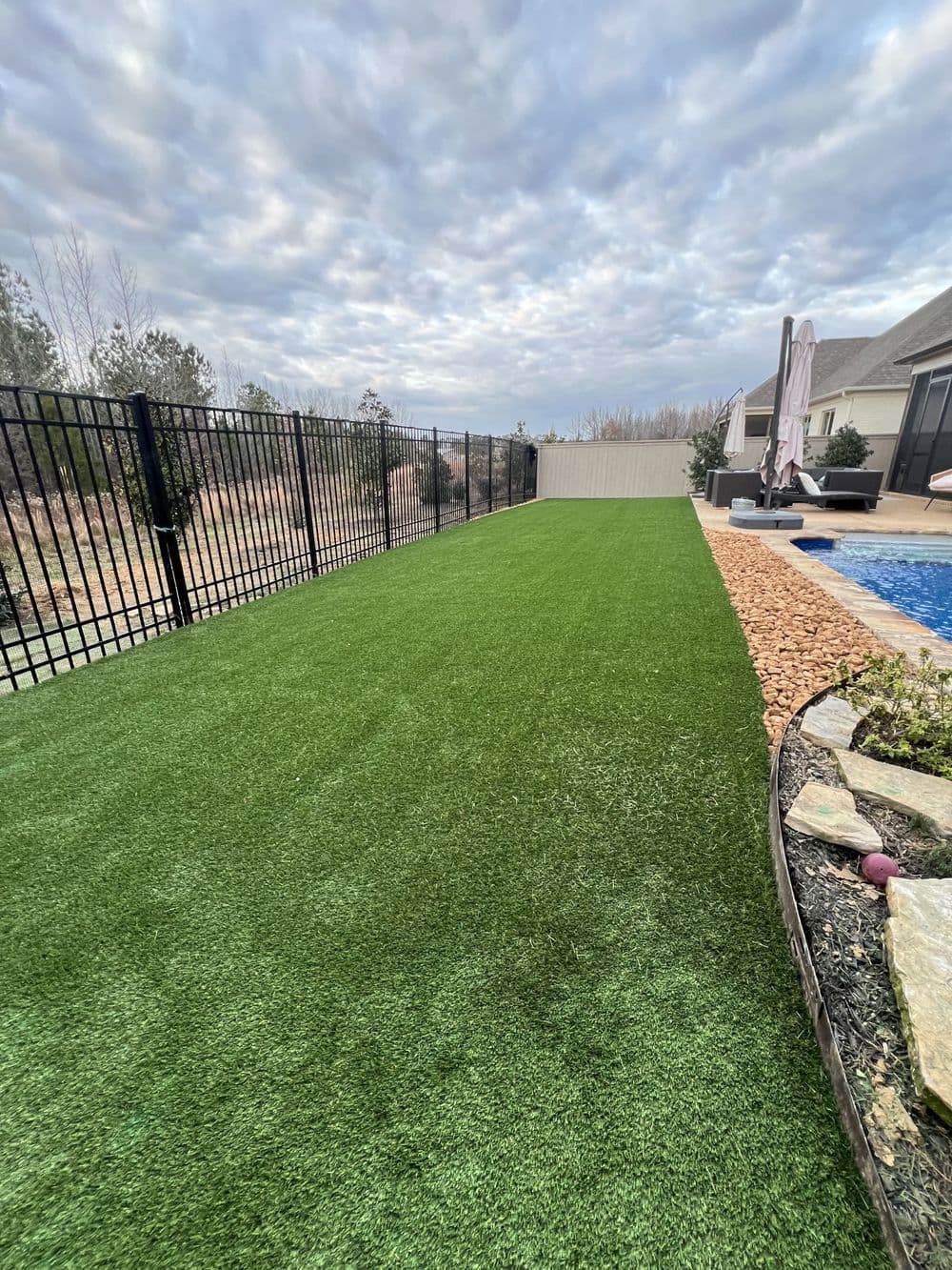 Lush green artificial lawn beside a pool with decorative rocks and a black fence.